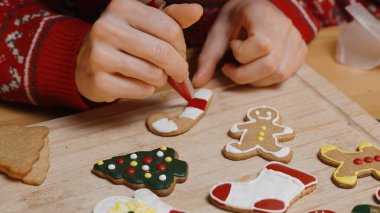 Kadın, Tahta Tahta Kurulda çeşitli şenlikli kurabiyelerle çevrelenmiş, Kırmızı ve Beyaz Buz Şekeriyle Şeker Çubuğu Kurabiyesi süslüyor..