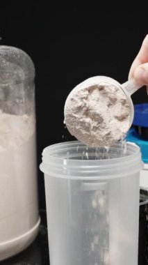 Dikey video. Sporcu, protein tozunu şişeye boşaltıyor, egzersiz sonrası beslenme artışı için karıştırmaya hazır. Yavaş çekim.
