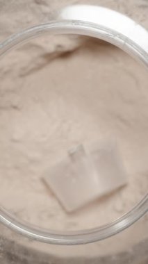 Dikey video. Sporcu, büyük bir konteynırdan Protein tozu topluyor ve egzersiz sonrası sarsıntıya eklemeye hazırlanıyor. Yavaş çekim.