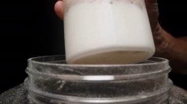 Bir el şeffaf bir Shaker bardağını bir kabın üzerinde tutar ve bir kepçeden bardağa Protein tozu döker..