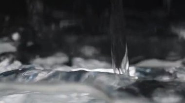 Bir su akıntısı bir kabın içine dökülür, yüzeyde dalgalanmalar ve kabarcıklar yaratır, karanlık bir arka plana yakın çekim detaylarıyla yakalanır. Yavaş Hareket