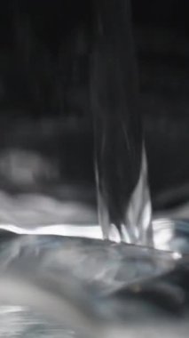 Dikey video. Bir su akıntısı bir kabın içine dökülür, yüzeyde dalgalanmalar ve kabarcıklar yaratır, karanlık bir arka plana yakın çekim detaylarıyla yakalanır. Yavaş Hareket