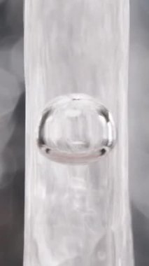 Dikey video. Tek bir hava kabarcığı, şeffaf bir tüple çekilen temiz bir su akımının merkezinde asılı duruyor ve makro detaylardaki dinamik akışı vurguluyor. Yavaş Hareket