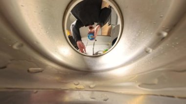 Metalik bir lavabonun içinden Macro zoom görüntüsü, bir adamın bardağa su doldurup içtiğini gösteriyor. Su damlacıkları lavabonun yüzeyinde görülebiliyor, bu da yakın bir perspektif yaratıyor. Suyun geri kalanını döküyor.
