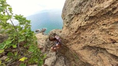 Bir kadın dikkatlice sarp kayaların arasına tırmanıyor, okyanus çok aşağıdayken. Engebeli uçurumlar kıyı şeridini dramatik bir şekilde gösteriyor..