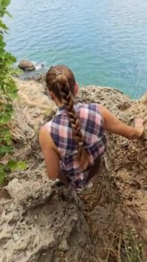 Dikey video. Bir kadın dikkatlice sarp kayaların arasına tırmanıyor, okyanus çok aşağıdayken. Engebeli uçurumlar kıyı şeridini dramatik bir şekilde gösteriyor..