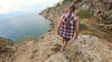 Bir kadın, sarp kayalıklar ve kuru bitki örtüsüyle çevrili, deniz kıyısındaki kayalık bir yoldan iner. Okyanus bulutlu gökyüzünün altında uzanıyor..