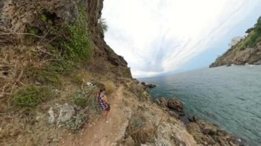 Bir kadın bir tarafında kayalıklar, diğer tarafında deniz olan kıyı şeridi boyunca yürür. Geniş açılı mercekle çekilen çekim geniş manzarayı yakalıyor..