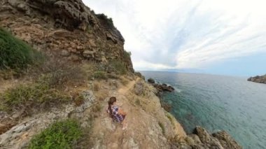 Bir kadın deniz kenarındaki dar bir uçurum boyunca yürür, bir tarafında yüksek uçurumlar vardır. Video geniş açılı mercek kullanılarak çekilir ve geniş manzarayı yakalar..