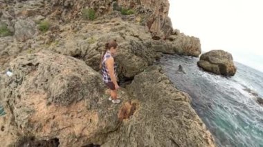 Bir kadın, sarp kayalıklar ve engebeli arazilerle çevrili, su kenarındaki büyük kayaların üzerinden tırmanıyor. Sahne geniş açıyla çekiliyor, zorlu manzarayı vurguluyor..