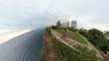 Bir erkek ve kadın okyanus manzaralı uçurum kenarında yürüyorlar. Sahne 360 derece açıyla çekilir, deniz ve çevreleyen yeşilliğin panoramik bir perspektifini sunar..