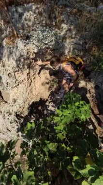 Dikey video. Bir kadın, aşağıdaki turkuaz suda bir tekneyle, dik kayalardan denize doğru tırmanıyor. Sahne geniş açılı bir perspektiften çekiliyor, sarp kayalıkları ve okyanus manzarasını yakalıyor..