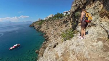 Bir kadın denizin üzerinde dar bir kıyı yolu boyunca yürüyor, uzaktaki bir tekneyle. Çekim geniş açılı mercekle, dramatik yüksekliği ve berrak suyu vurguluyor..