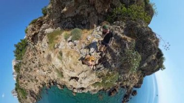 360 derecelik kamera ile bir adam ve bir kadın denizin üzerinde dar bir uçurum kenarında yürüyorlar. Video geniş açıyla çekiliyor. Dik düşüş ve aşağıdaki sudaki tekneyi gösteriyor..