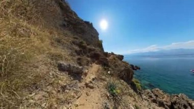 Bir kadın okyanusa doğru kayalık bir patika boyunca inişe devam ediyor, suda görünen bir tekneyle. Video birinci şahıs perspektifinden çekilmiştir, engebeli yolu vurgular..