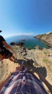 Dikey video. Bir adam kayalık bir sahil yolu boyunca yürür, denize doğru bakarken geriye bakar. Video birinci şahıs perspektifinden çekiliyor, aşağıdaki temiz suyu vurguluyor..