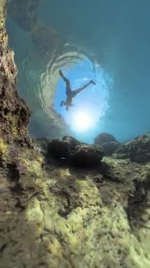 Dikey video. Bir Adam, bir Rocky Mağarası açılışı çerçevesinde Sunlight 'ın aktığı sualtında yüzer. Çekim geniş açılı bir açıdan çekiliyor, dramatik mağarayı ve berrak suyu vurguluyor..