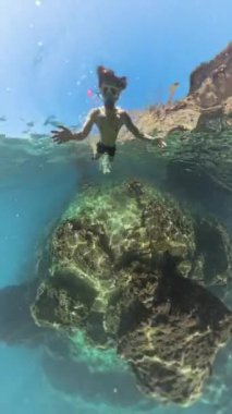 Dikey video. Bir adam büyük bir su altı kayasına doğru yüzüyor, şnorkel maskesi takarak uzanıyor. Geniş Açı, Su Yüzeyi 'ne Yakın Yakaladık..