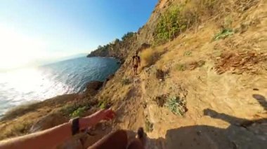 Bir Kadın, Kayalık Kıyı Yolu Boyunca Bir Erkeği Takip Ediyor, Gökyüzündeki Güneş Alçak, Kayalıklarda ve Altındaki Parlak Deniz 'de Altın Bir Parıltı Yaratıyor.