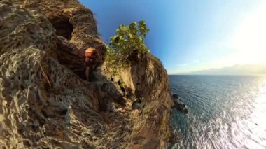 Bir adam ve bir kadın sarp bir uçuruma tırmanıyor, Etrafı engebeli kayalarla çevrili, engin, parıldayan okyanuslarla dolu.