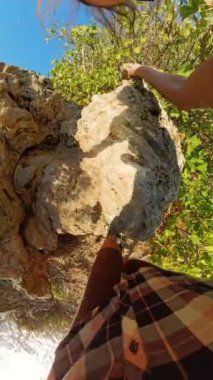 Dikey video. Kayalık Kayalıklara tırmanan bir kadının ilk insan görüntüsü. Altında köpüklü okyanus ve parlak güneş ışığı sudan yansıyor..