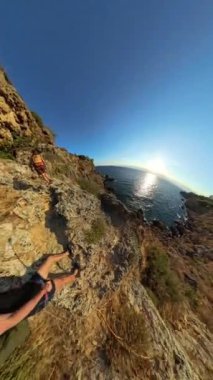 Dikey video. Bir çift, uçurum boyunca sarp kayalıklara tırmanıyor, okyanusun üzerinde yükseliyor, Güneş batmaya başlarken tepeye doğru ilerliyor..