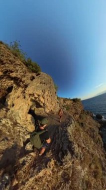 Dikey video. Okyanusa bakan bir uçurumun kenarındaki küçük bir mağarada dinlenen iki kişi küresel bir panorama içinde yakalandı..