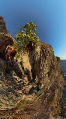 Dikey video. Bir adam ve bir kadın sarp bir uçuruma tırmanıyor, Etrafı engebeli kayalarla çevrili, engin, parıldayan okyanuslarla dolu.