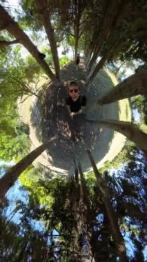 Dikey video. Ormanda küçük bir gezegen etkisi, merkezde uzun ağaçlarla çevrili bir insan, dairesel dünya benzeri bir illüzyon yaratıyor, doğal çevrenin gerçeküstü bir görüntüsünü sunuyor..