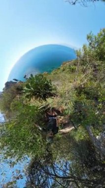 Dikey video. Kaktüslere yakın, okyanusa bakan, kıyı şeridinin çarpıcı bir şekilde kıvrıldığı, kıyı manzarası ve gökyüzünün küresel, dalgın bir görüntüsünü yaratan bir adamın küçük gezegen etkisi..