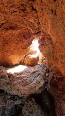 Dikey video. Dolly, gün ışığıyla aydınlatılan kayalık bir mağaranın içinden bakıyor. Kayaların ılık turuncu tonlarını aydınlatıyor. Işık bir açıklıktan sızıyor ve gizemli bir doğal geçit oluşturuyor..