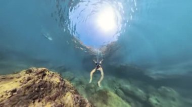 Snorkeler su altında büyük bir kayaya tırmanıyor, güneş ışığı yüzeye ulaşıyor, kayanın canlı dokusunu ve çevresindeki deniz ortamını vurguluyor..