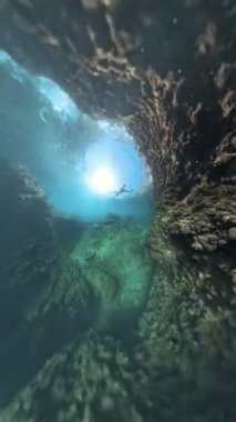 Dikey video. 360 derecelik Panorama Snorkeler, kayalık oluşumların yakınında süzülen dalgıç, berrak suda parlayan güneş ışığıyla büyük bir su altı kayasının çevresini keşfediyor..