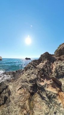 Dikey video. Rocky Cliffs 'in ön planda olduğu sahil sahnesi, dalgalar aşağı düşüyor. Güneş yüksektir ve canlı bir Açı etkisi yaratır. Okyanus ve Kayaları yakalar..