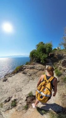 Dikey video. Rocky Cliffside Patikası 'nda yürüyen bir kadının geniş açılı görüntüsü, soldaki parlak güneşin altında parlayan okyanus, güzel bir sahil manzarası yaratıyor..