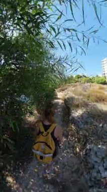 Dikey video. Sırt çantalı bir kadın uzun kuru otlar ve çalılar arasında dar bir yolda yürüyor ve binalar uzaktan görülebiliyor. Sahne geniş açılı bir efektle çekilir.
