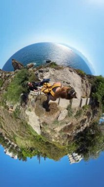 Dikey video. Bir kıyı manzarasının küçük gezegeni etkisi. Bir adam ve kadın okyanusa tepeden bakan bir patikada yürüyor ve manzaranın küresel bir görüntüsünü yaratıyor..