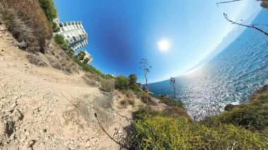 Sahil Yolu 'nun küresel panoraması. Önünde engebeli arazi ve çalılıklar var. Okyanus güneşin altında parıldıyor ve binalar bu etkiye bağlı olarak kıvrımlı görünüyor..