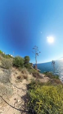 Dikey video. Sahil Yolu 'nun küresel panoraması. Önünde engebeli arazi ve çalılıklar var. Okyanus güneşin altında parıldıyor ve binalar bu etkiye bağlı olarak kıvrımlı görünüyor..