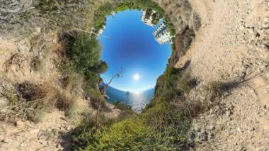 Kıyı yolunun küresel panoraması, ön planda kuru bir Rocky arazisi var. Bitki örtüsü ve binalar kıvrılarak uzak okyanusu parlak bir güneş altında çerçeveler..