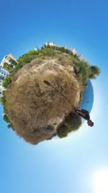 Dikey video. Küçük Gezegen 360 efekti, bir adamın Rocky Arazisinde durduğunu, çevresindeki bitki örtüsüyle okyanus ve binaların, açık mavi gökyüzü altında küre ufkunun etrafında kıvrıldığını gösteriyor..