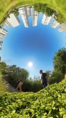 Dikey video. Gezegen Deliği etkisi, uzun yeşil çalılarla çevrili bir yolda yürüyen bir adam ve kadının etrafında dairesel bir manzara oluşturan binalar ve açık mavi gökyüzü..