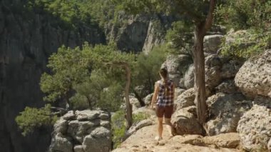 Bir kadın, kayalar ve ağaçlarla çevrili engebeli bir patikada yürüyor. Huzurlu bir Kanyon ortamında uçurumun kenarına yaklaşıyor. Arka planda sarp kayalıklar görünüyor..