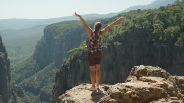 Bir kadın, kollarını zaferle kaldırarak uçurumun kenarında duruyor. Kanyon 'un nefes kesici manzarasını kucaklıyor. Geniş, dağlık manzara onun arkasında uzanıyor..
