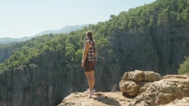 Bir kadın kayalık bir uçurumun kenarında duruyor, kolları geniş, geniş Kanyon manzarasına bakıyor. Engebeli arazi ve yeşil orman kontrastı açık gökyüzü ile.