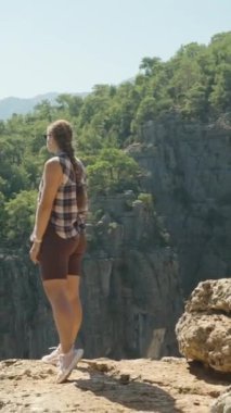 Dikey video. Bir kadın kayalık bir uçurumun kenarında duruyor, kolları geniş, geniş Kanyon manzarasına bakıyor. Engebeli arazi ve yeşil orman kontrastı açık gökyüzü ile.