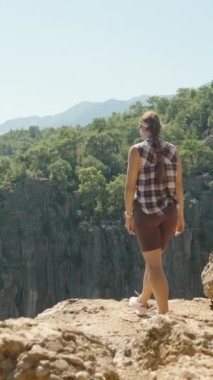 Dikey video. Bir kadın kollarını açıp kayalık bir uçurumun kenarında duruyor. Geniş kanyona ve parlak güneşin altındaki orman dağlarına bakıyor..