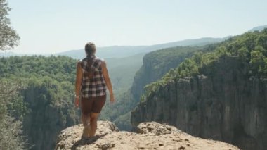 Bir kadın kayalık bir çıkıntıda duruyor, kolları açık, Kanyon 'un ve etrafındaki vahşi doğanın güzelliğini kucaklıyor, spor ayakkabılar giyiyor ve uzak kayalıkların manzarasını seyrediyor..
