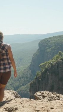 Dikey video. Bir kadın kayalık bir çıkıntıda duruyor, kolları açık, Kanyon 'un ve etrafındaki vahşi doğanın güzelliğini kucaklıyor, spor ayakkabılar giyiyor ve uzak kayalıkların manzarasını seyrediyor..