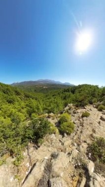 Dikey video. Kayalık bir patika, arka planda dağlar olan sık bir ormana bakar. Geniş açı perspektifi parlak bir gökyüzünün altındaki engebeli araziyi ve geniş yeşilliği vurgular..